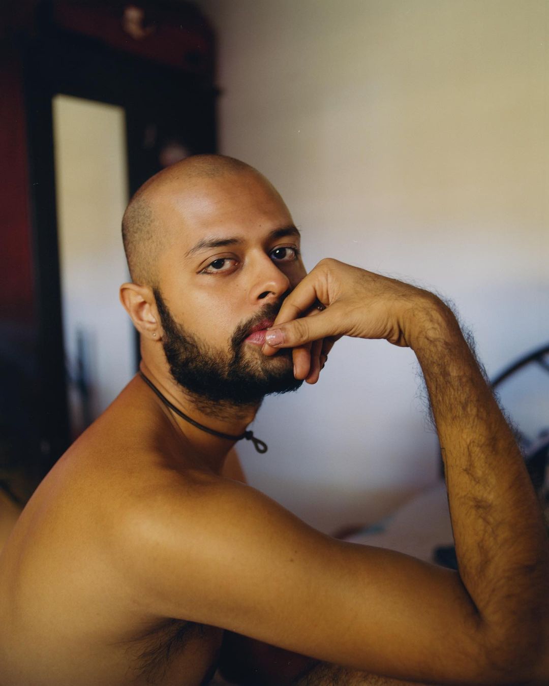 Portrait of a shirtless, bald man with a beard indoors, gazing at the camera with a neutral expression and his fingers near his mouth.
