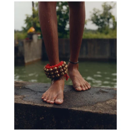 Close-up of a young person standing barefoot near a body of water, wearing traditional ankle bells and a bracelet in a natural setting.