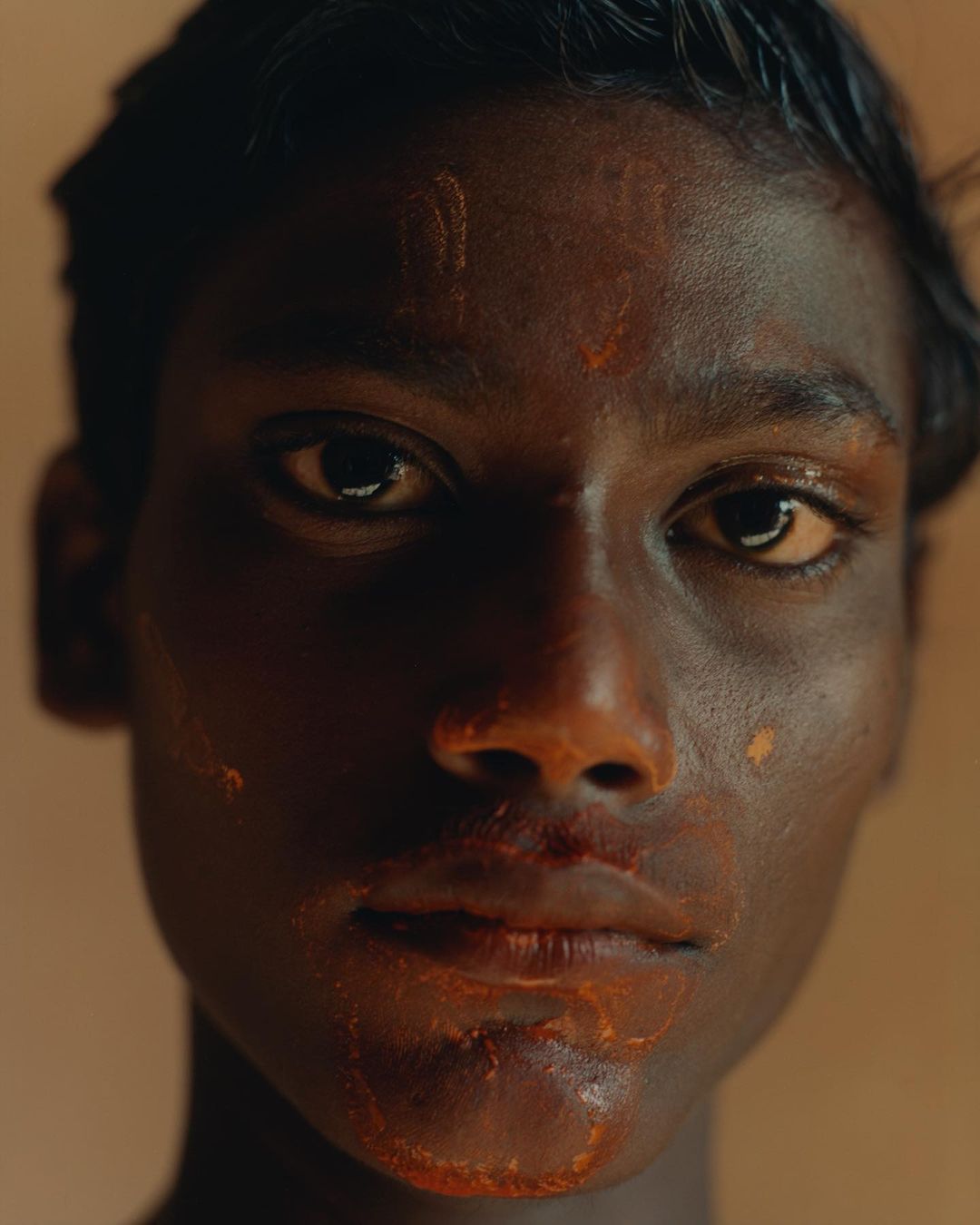 Close-up portrait of a young adult with dark skin, adorned with orange paint on their face, under soft lighting and earth-toned backdrop.