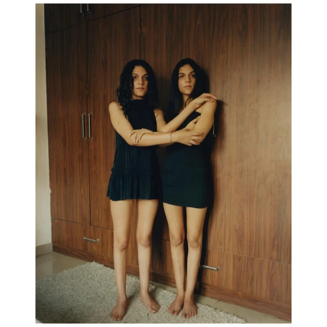 Two young adult women stand barefoot side-by-side in front of a wooden wardrobe wearing dark dresses, in an indoor setting on a carpet.