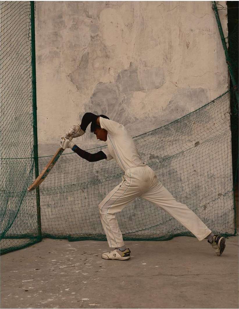 Male cricket player in white uniform hits ball with bat during practice at outdoor training facility with net and weathered wall.