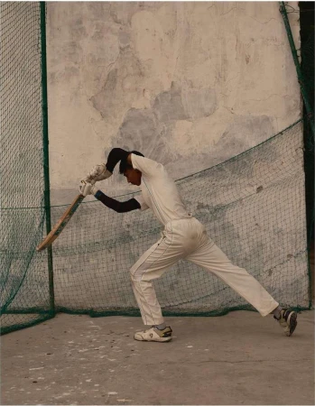 Male cricket player in white uniform hits ball with bat during practice at outdoor training facility with net and weathered wall.