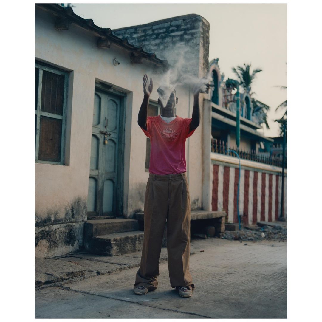 Man throwing dust into the air outside a building. He is wearing a t-shirt and pants, and the shot is an eye-level medium shot.