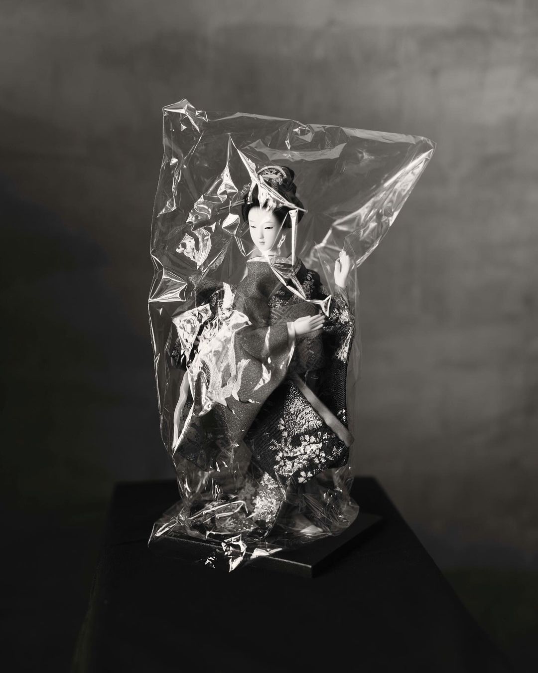 Monochromatic photo of a Japanese doll in traditional kimono dress displayed in a plastic bag, on a dark stand, against a grey background.