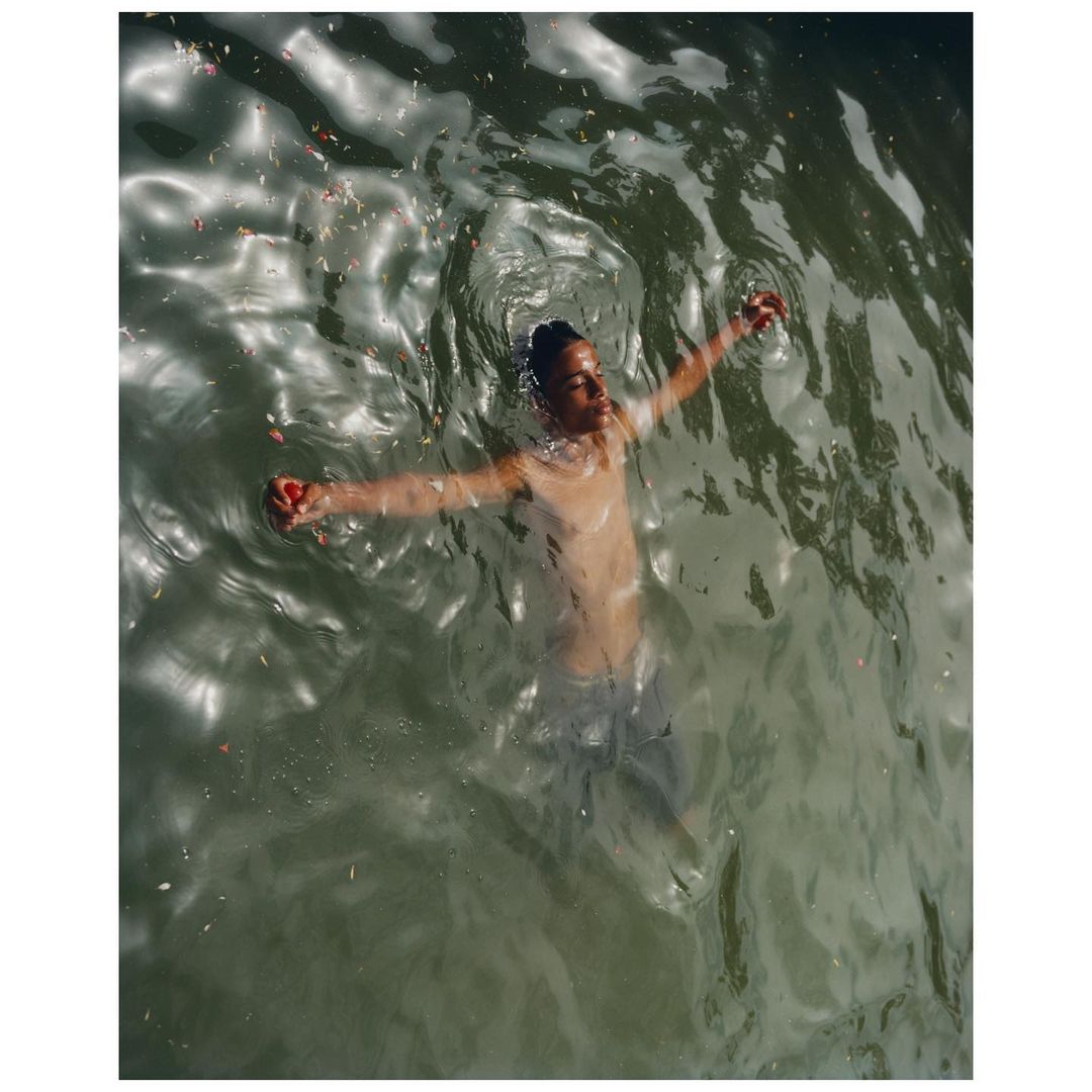 Young adult floats on their back in rippling greenish water with arms outstretched, serene outdoor scene in natural light.