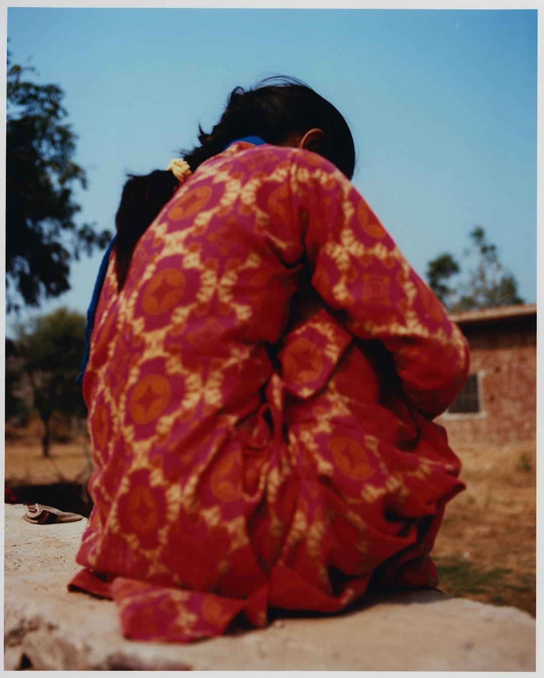 Child in patterned red dress sits outside with blurred building and blue sky in background, captured in a documentary style.
