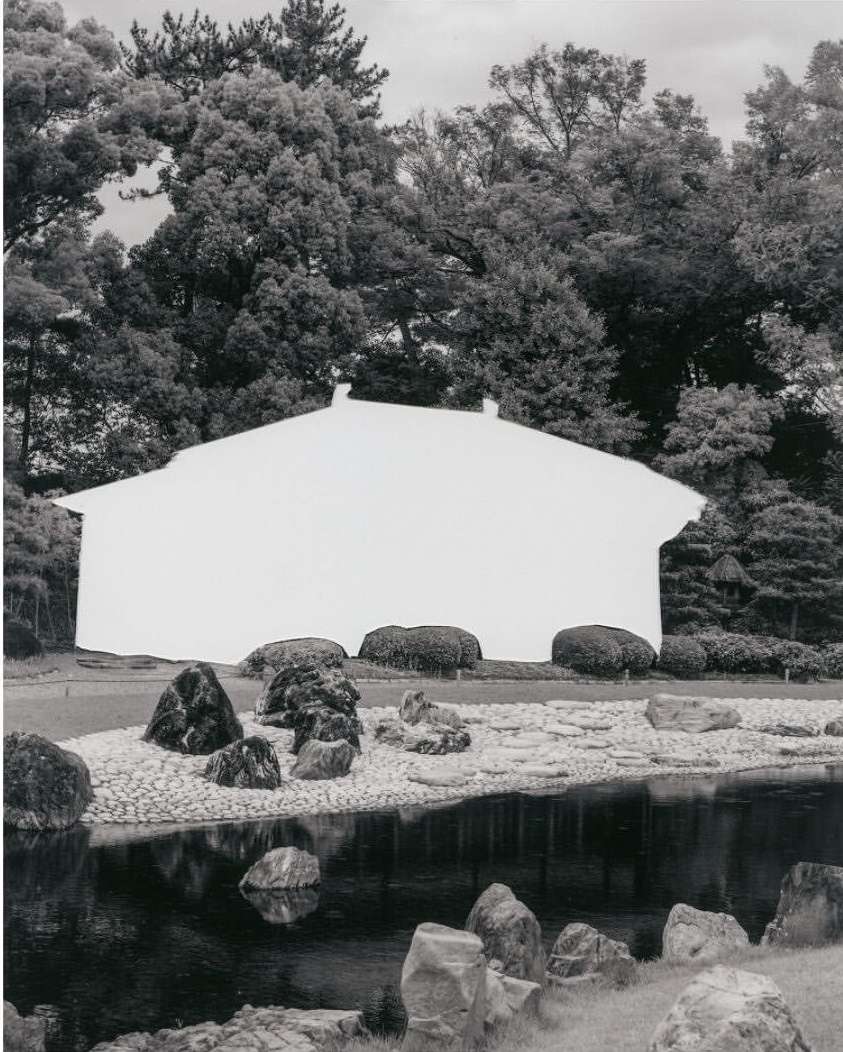 Monochrome photograph of white abstract facade with dark roof against trees in a landscape with rocks and water creating an artistic image.