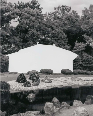 Monochrome photograph of white abstract facade with dark roof against trees in a landscape with rocks and water creating an artistic image.