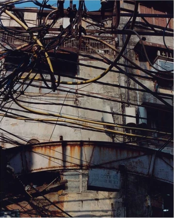 Exterior of an old building with peeling paint and rust, crisscrossed by a dense network of electrical wires and signage in daylight.