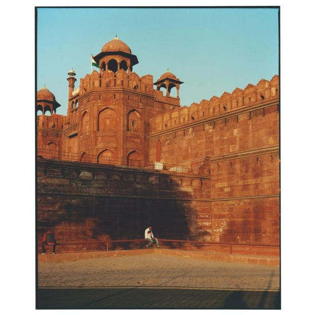 The Red Fort of India with its walls and domes, sits under a clear sky. A person sits in the foreground, adding scale to the monument.