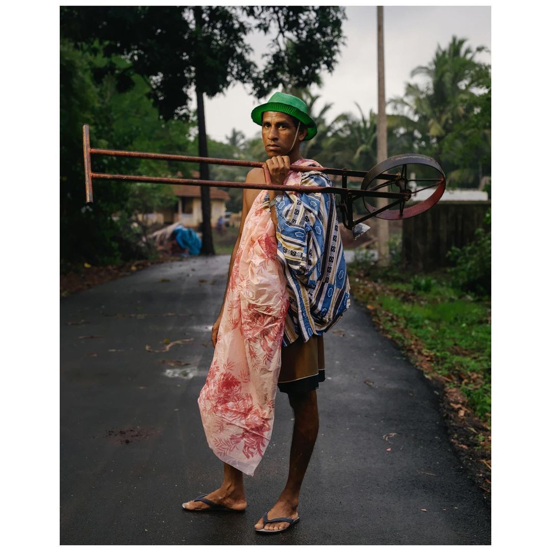 A man stands on a wet road carrying a metal frame and patterned textiles, with lush green trees and foliage surrounding him in this candid scene.