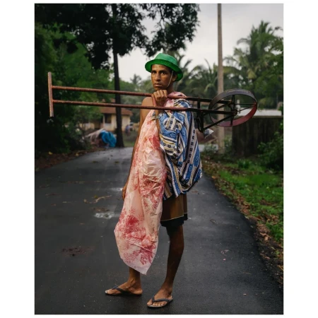 A man stands on a wet road carrying a metal frame and patterned textiles, with lush green trees and foliage surrounding him in this candid scene.