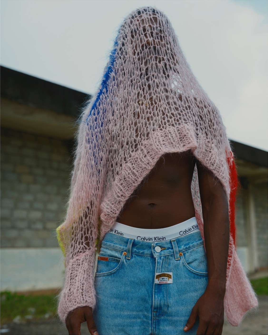A dark-skinned human stands outdoors wearing denim jeans, Calvin Klein underwear, and a multi-colored, see-through woven shawl covering the head.