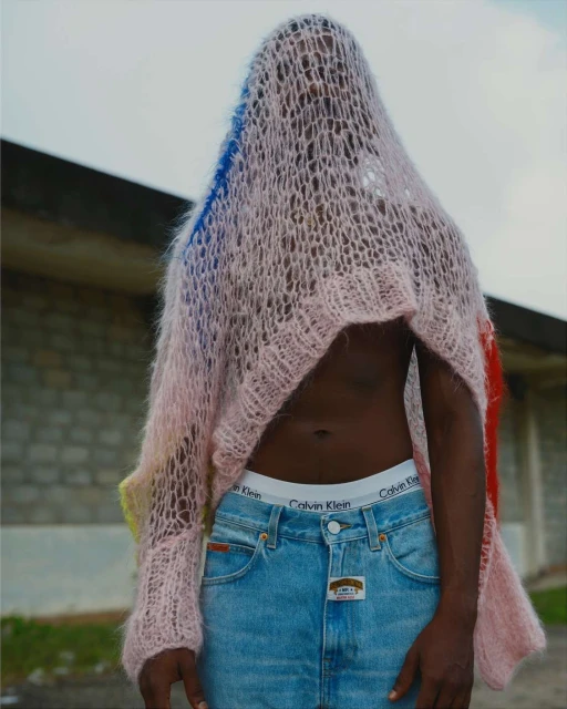 A dark-skinned human stands outdoors wearing denim jeans, Calvin Klein underwear, and a multi-colored, see-through woven shawl covering the head.