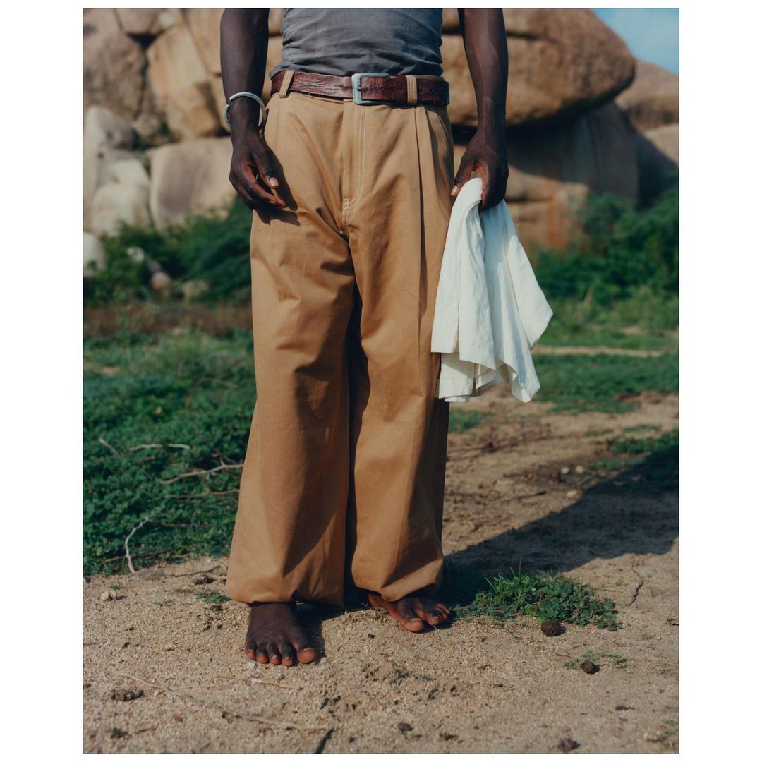A person stands barefoot in tan pants and a gray tank top, holding a white cloth in a natural, sunny outdoor setting.