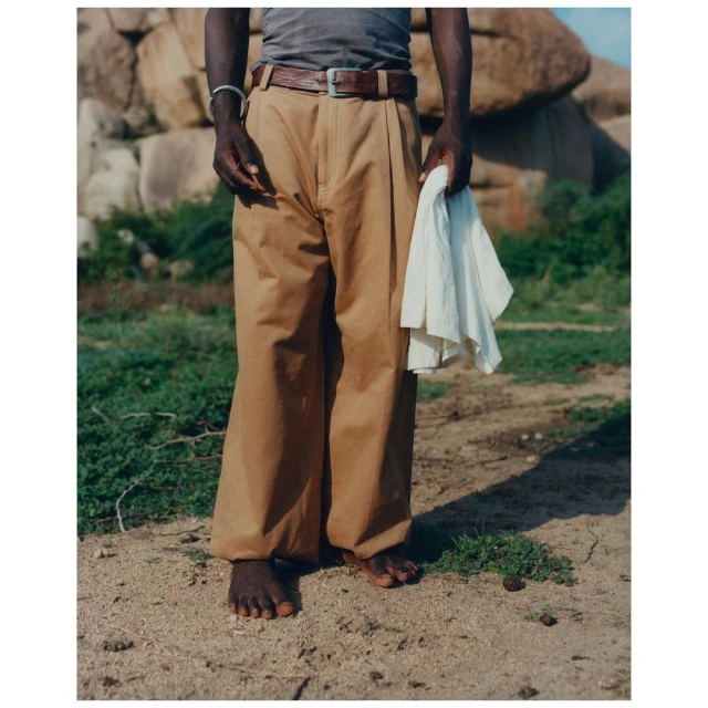 A person stands barefoot in tan pants and a gray tank top, holding a white cloth in a natural, sunny outdoor setting.