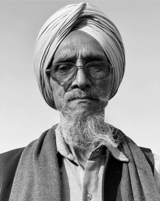 Portrait of an elderly man wearing a turban and glasses, with a distinguished beard and a thoughtful expression, against a bright sky.
