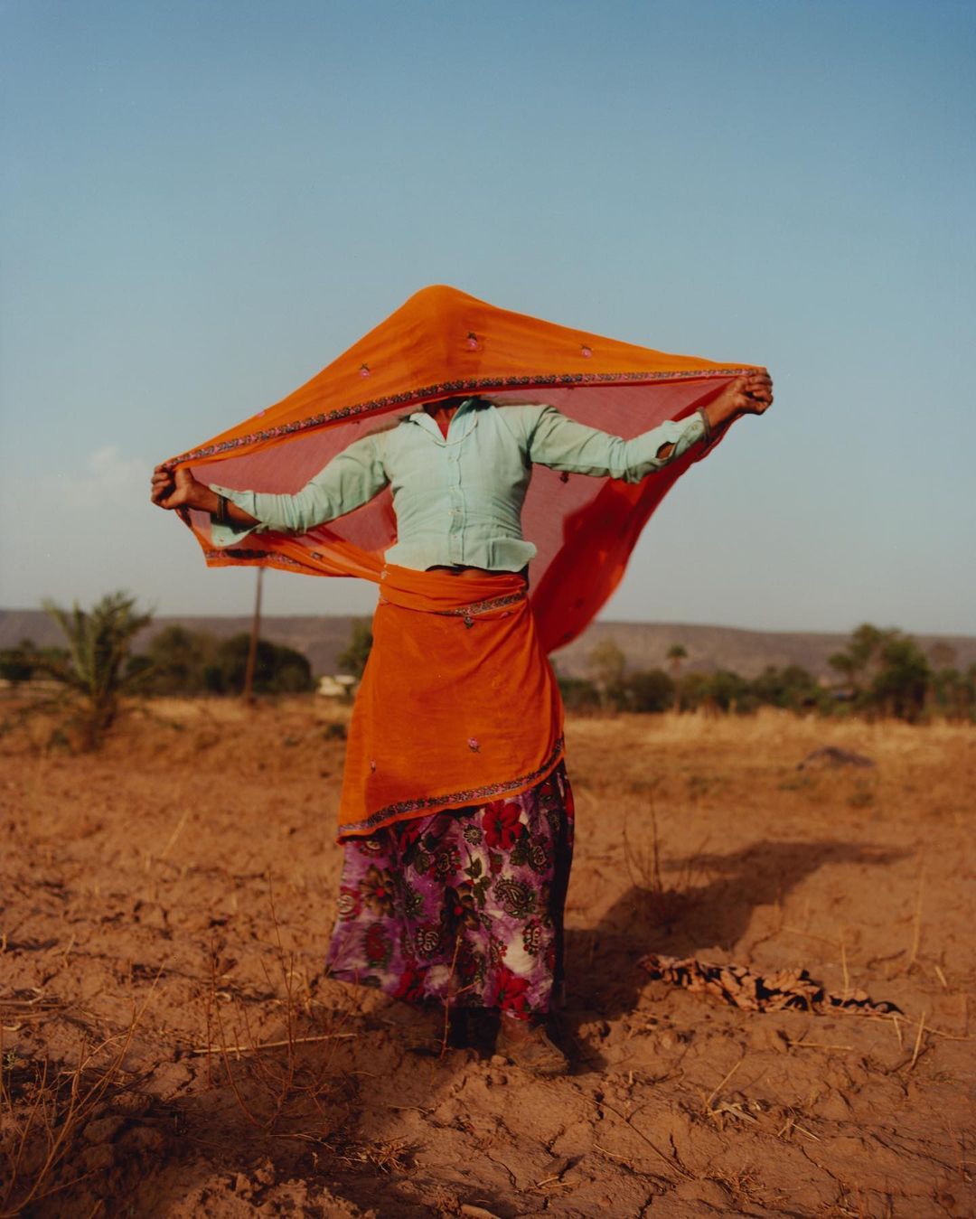 Woman in an orange sari stands in a field with a rural landscape, under a clear sky, and vibrant colors, creating a sense of place.