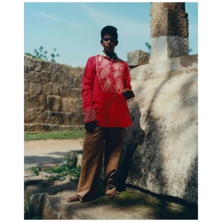 A young person stands outdoors next to a stone structure, wearing a red, patterned tunic and brown pants against a bright, daytime sky.