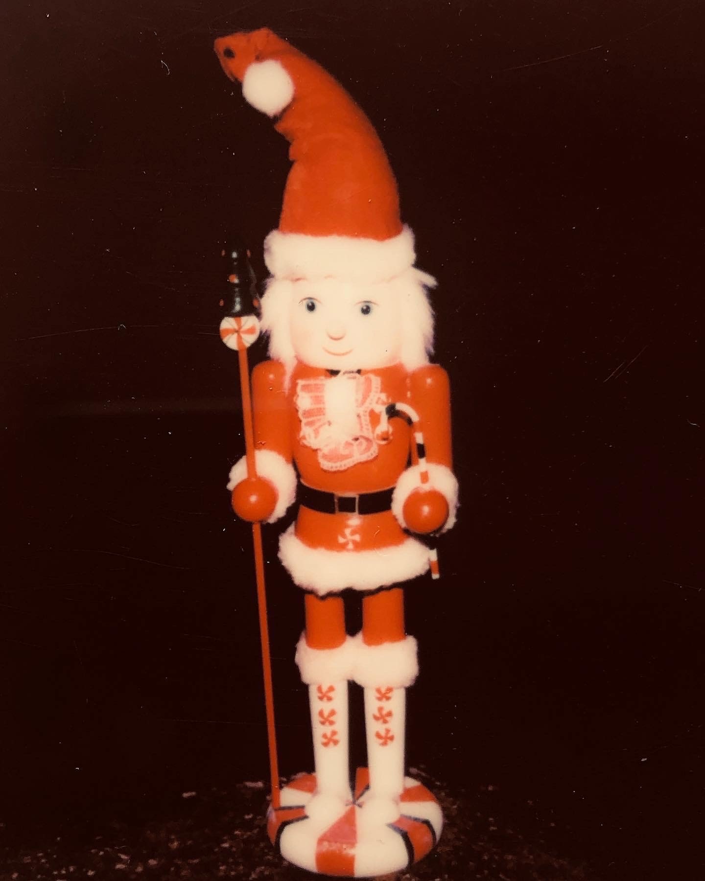 Vintage photograph of a toy nutcracker figure, with red and white holiday-themed colors against a dark background, in portrait orientation.