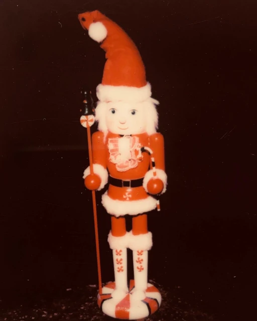 Vintage photograph of a toy nutcracker figure, with red and white holiday-themed colors against a dark background, in portrait orientation.