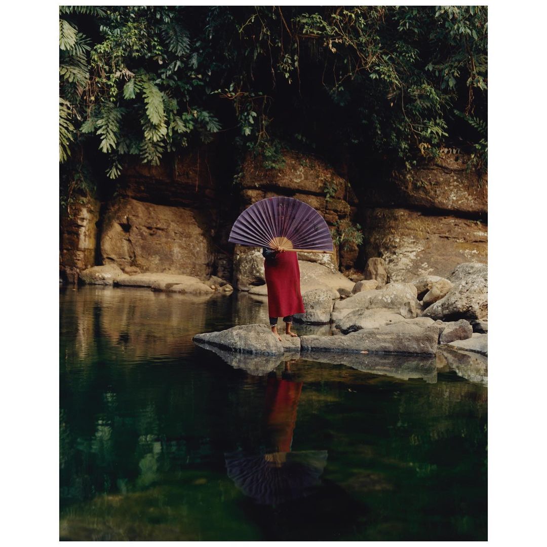 Person in red robe stands on river rocks with folded fan, reflected in water, surrounded by lush ferns and a rock cliff backdrop.
