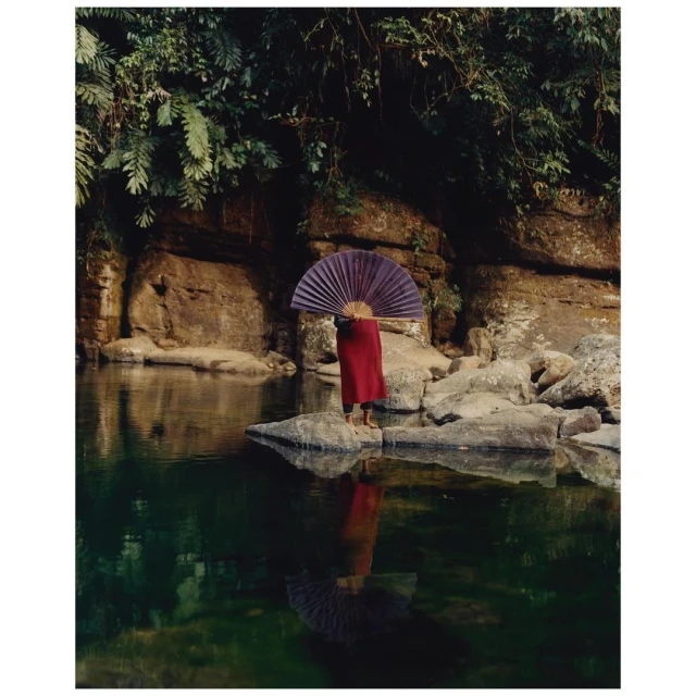 Person in red robe stands on river rocks with folded fan, reflected in water, surrounded by lush ferns and a rock cliff backdrop.