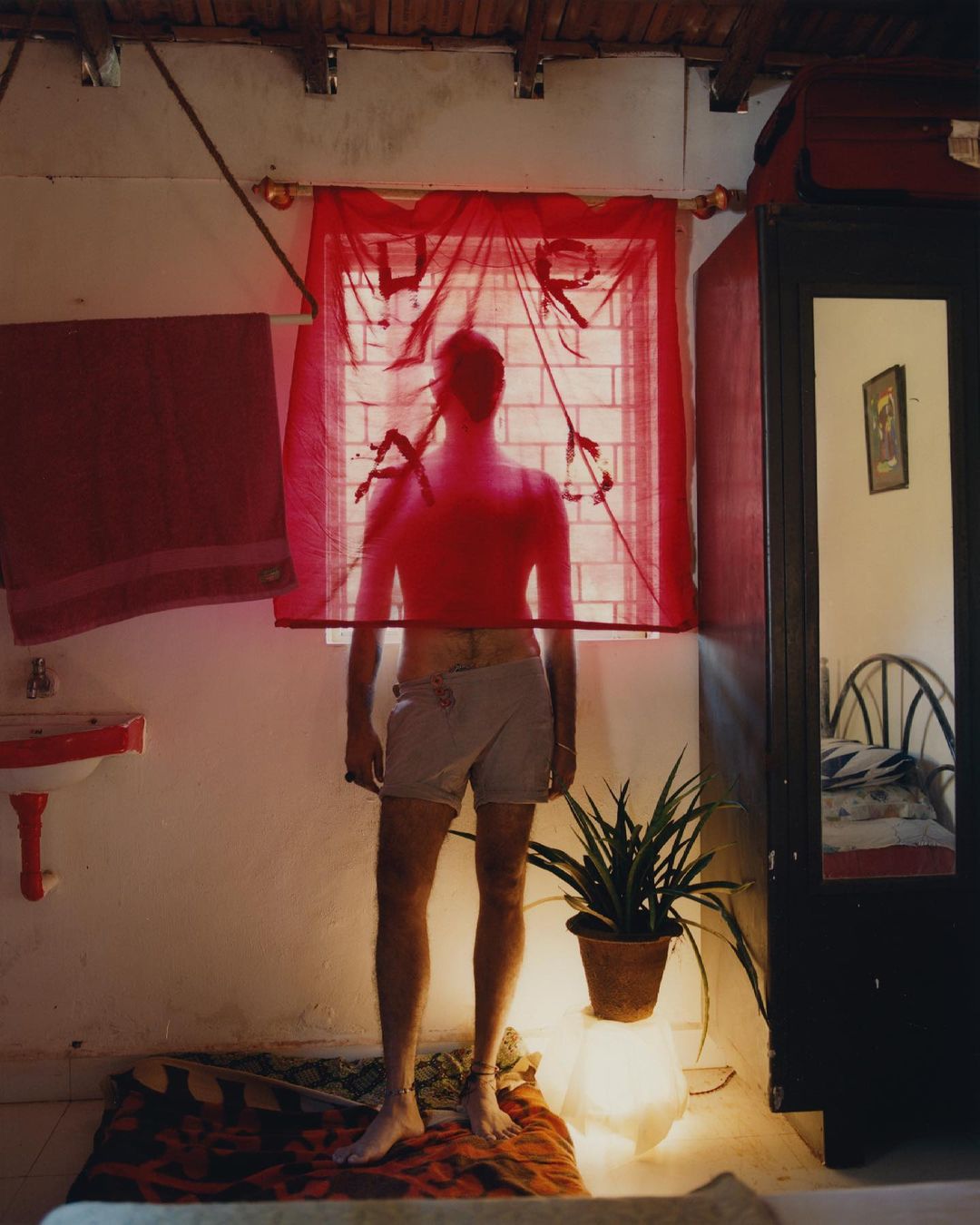 Back view of shirtless adult male standing indoors with light skin, wearing shorts, in front of window with a red transparent curtain.
