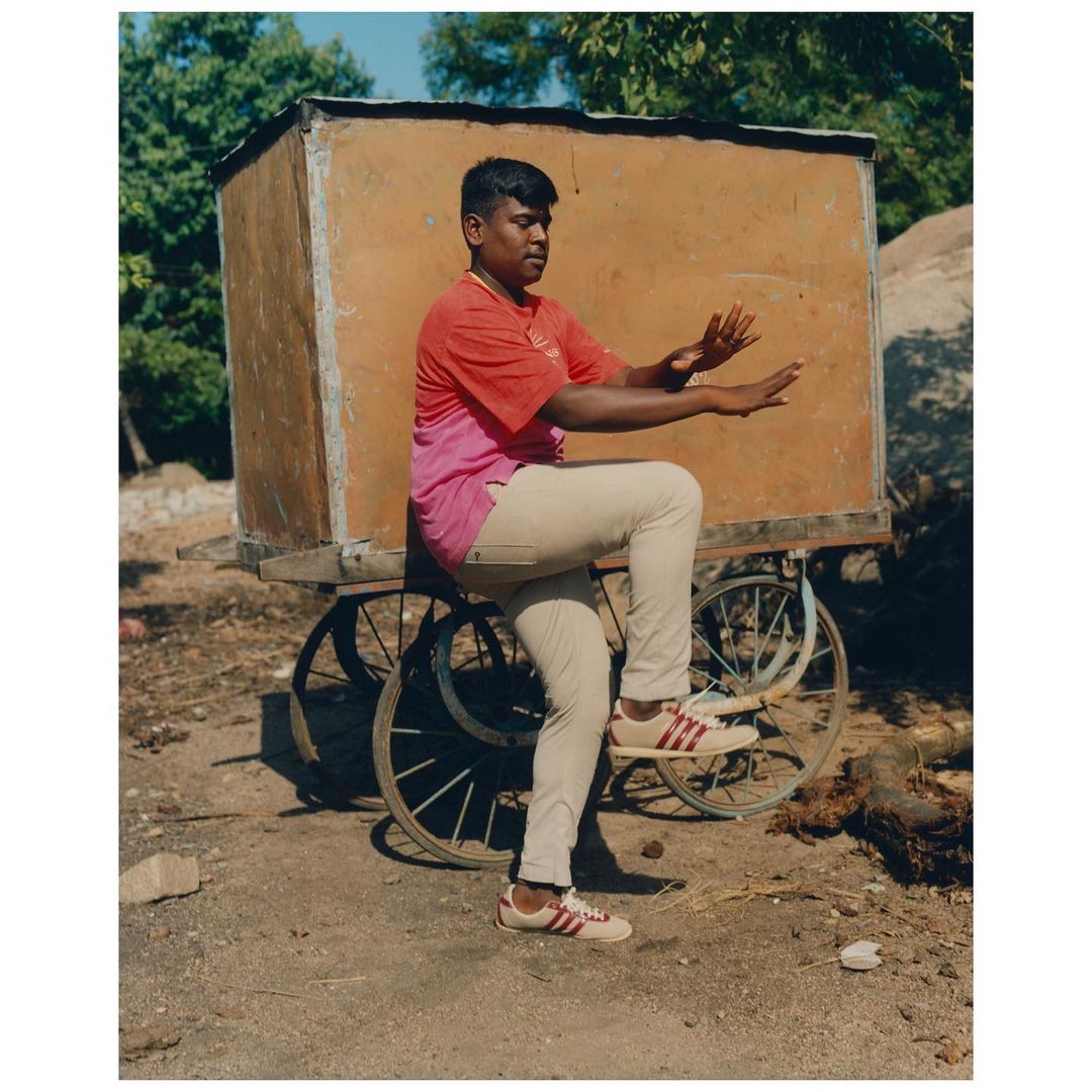 Young man balancing on a wheeled cart outdoors in sunlight, wearing jeans, sneakers, and a two-tone t-shirt, with trees in background.