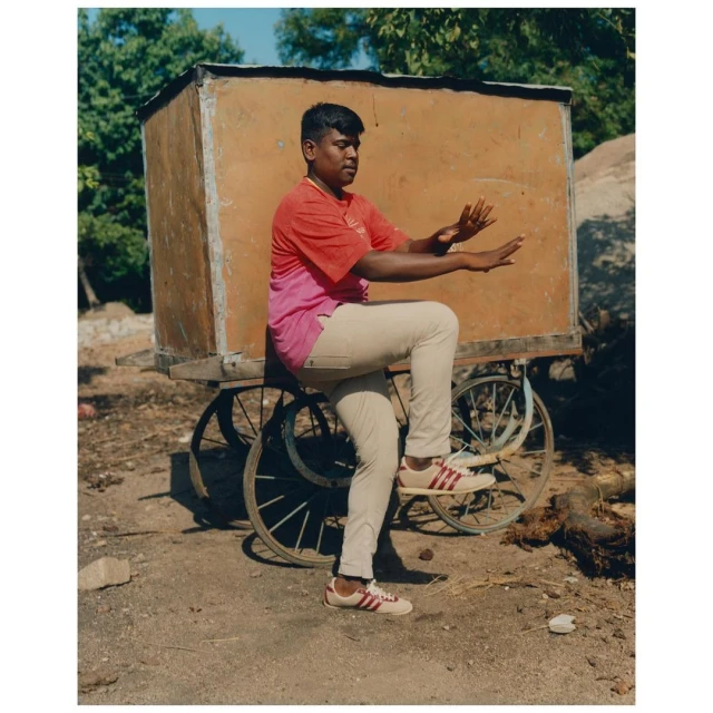 Young man balancing on a wheeled cart outdoors in sunlight, wearing jeans, sneakers, and a two-tone t-shirt, with trees in background.