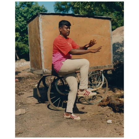 Young man balancing on a wheeled cart outdoors in sunlight, wearing jeans, sneakers, and a two-tone t-shirt, with trees in background.