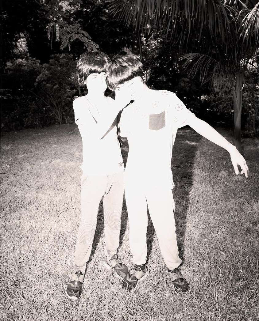 Two young boys standing closely together outdoors on a grassy lawn with trees in the background, captured in high-contrast monochrome.