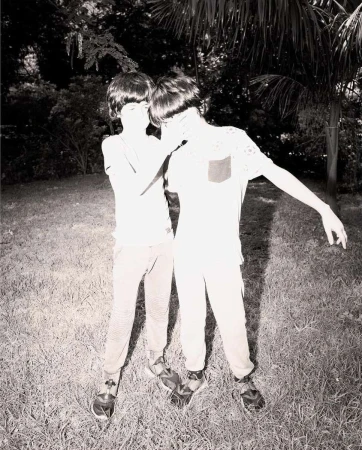 Two young boys standing closely together outdoors on a grassy lawn with trees in the background, captured in high-contrast monochrome.