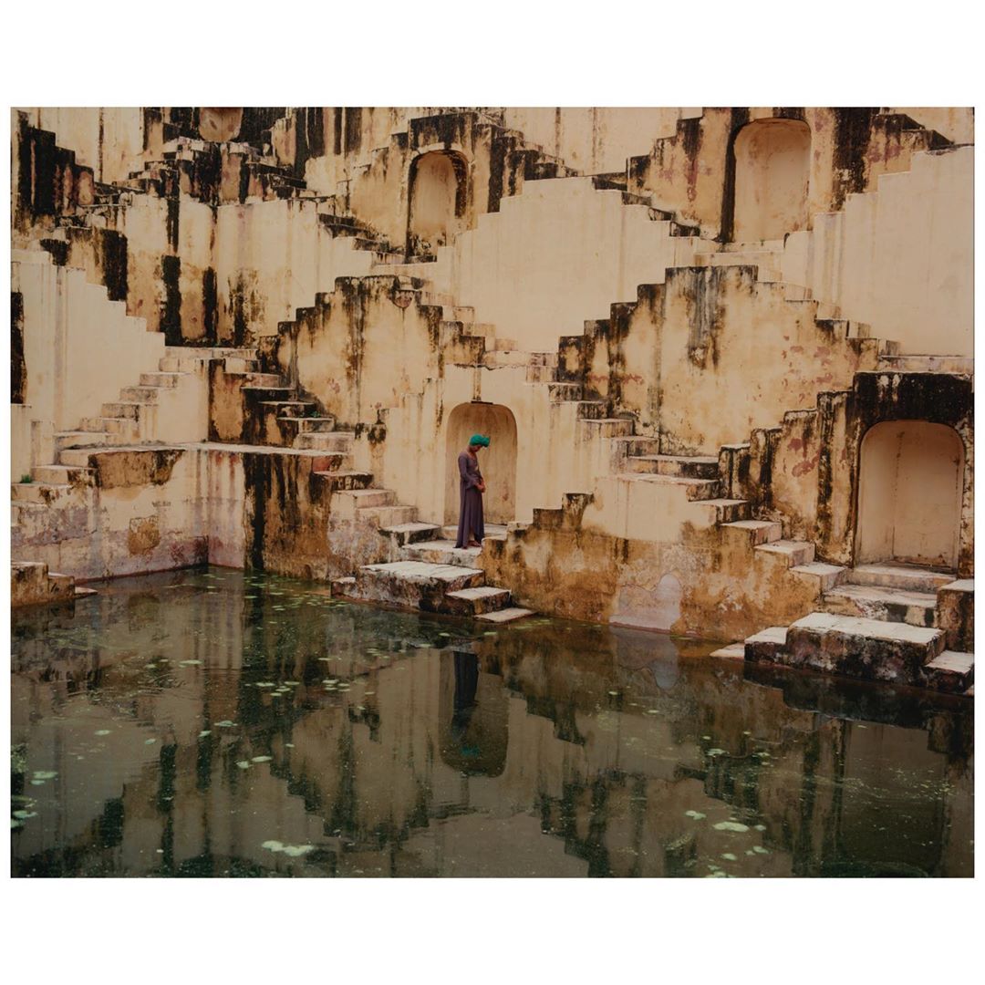 A person wearing a turban stands on the steps of the ancient Chand Baori stepwell in Rajasthan, India; water reflects the architecture.