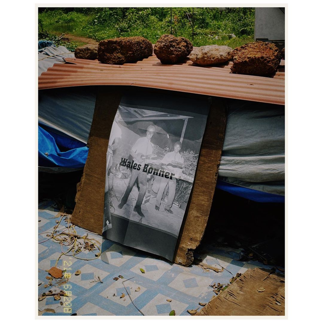 Outdoor display of a black and white 'Wales Bonner' poster leaned against wooden supports with rocks placed on top of a rusty metal sheet.