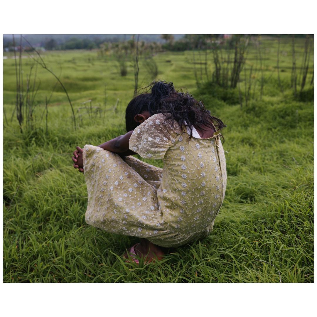 Child in a field wearing a dress, crouching with head bowed. Rural outdoor scene in a grassy field with blurred background.