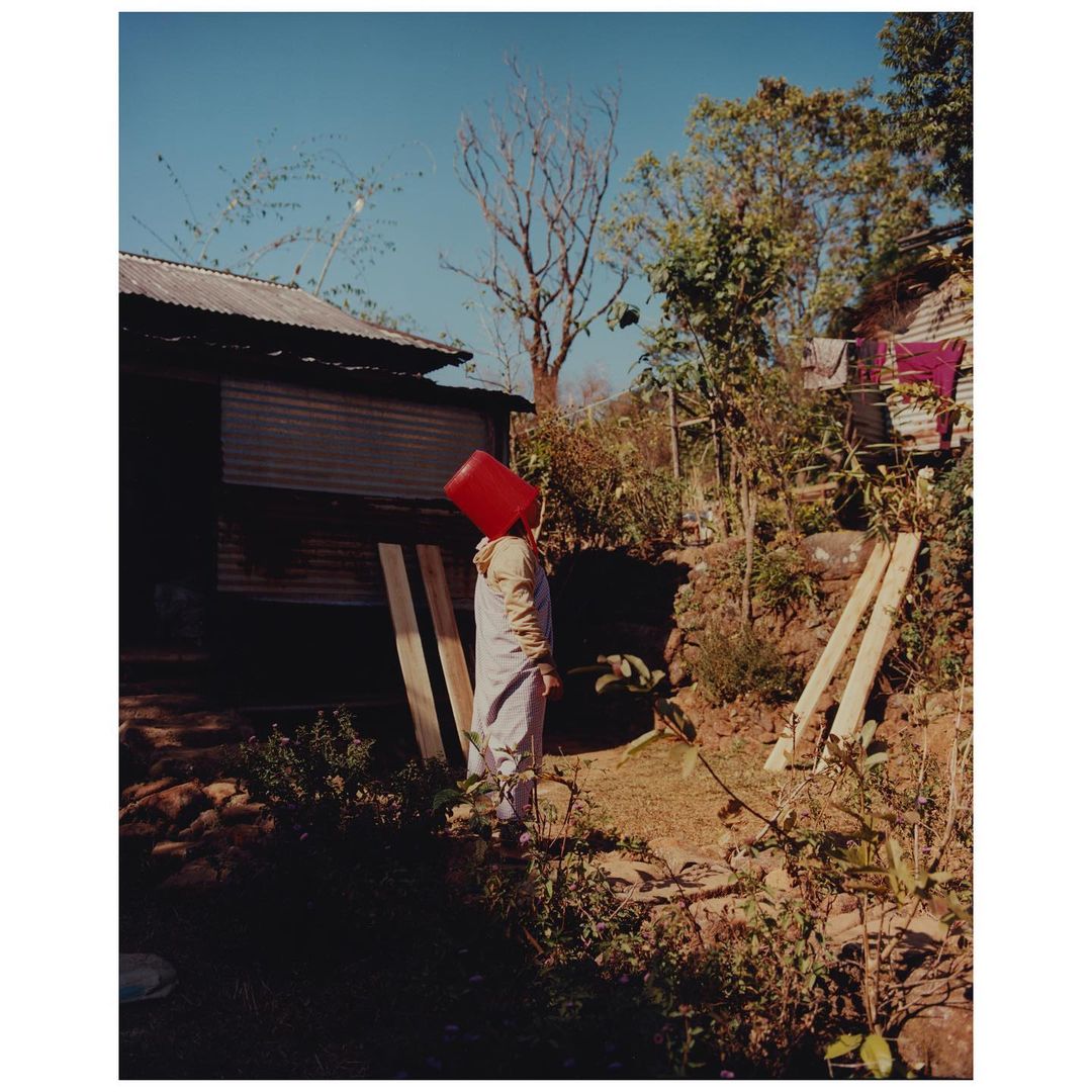 Person with red bucket on head stands in a yard with trees and a building. The sky is light blue, and the scene is outdoors.