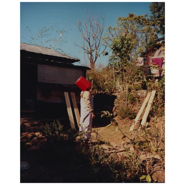 Person with red bucket on head stands in a yard with trees and a building. The sky is light blue, and the scene is outdoors.