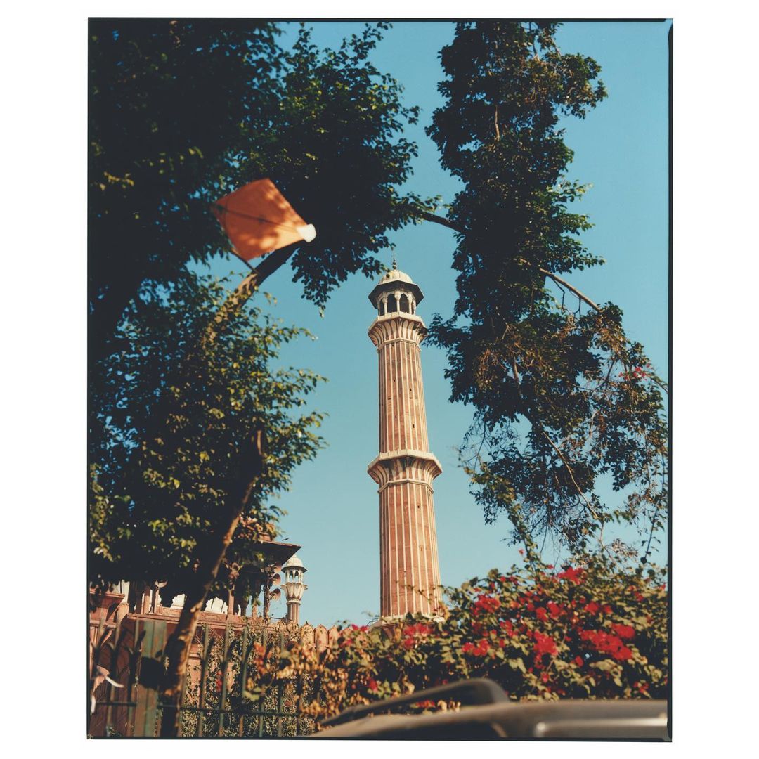 Tall minaret tower viewed from below, framed by green trees and red flowers against a clear blue sky on a sunny day.