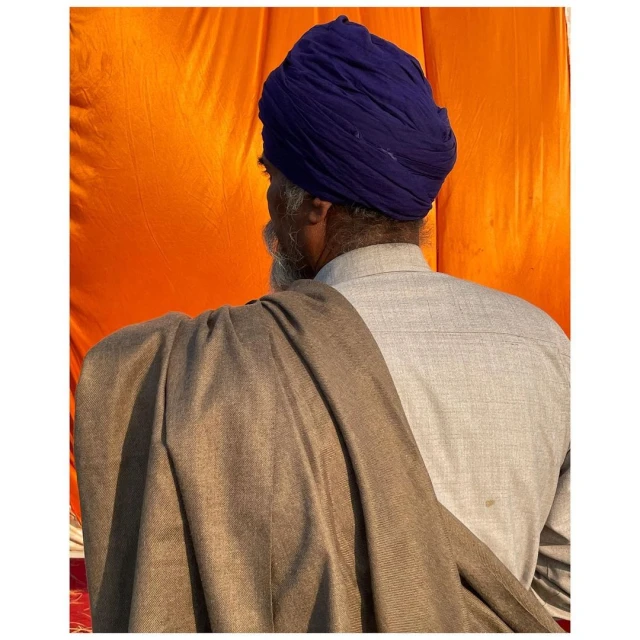Elderly Sikh man in blue turban and grey shirt with shawl. Head and shoulders from behind against a bright orange background.