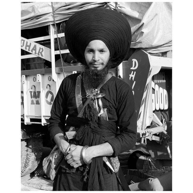 Monochrome portrait of a Sikh man with a large turban, beard, and traditional attire standing against a truck backdrop in daylight.