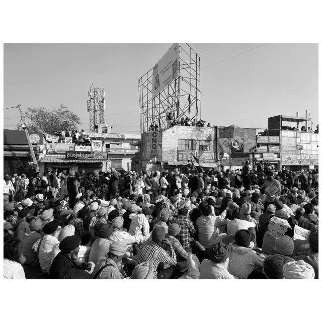 Large crowd of people gathered in an outdoor urban environment, some seated, others standing, buildings and a billboard in the background.