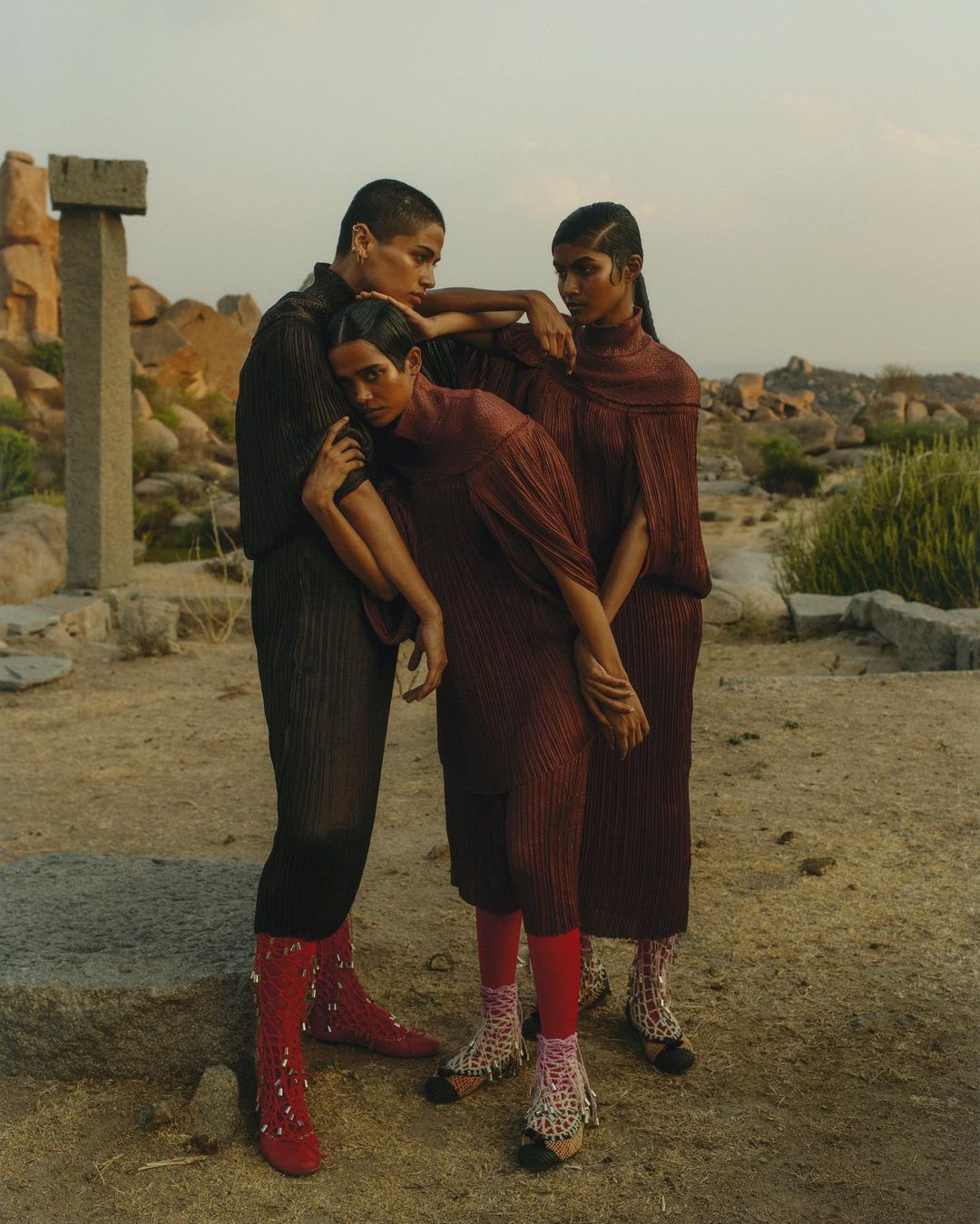 Three models in matching pleated tunics and red tights pose in a sunny outdoor location with scattered rocks and stone ruins.