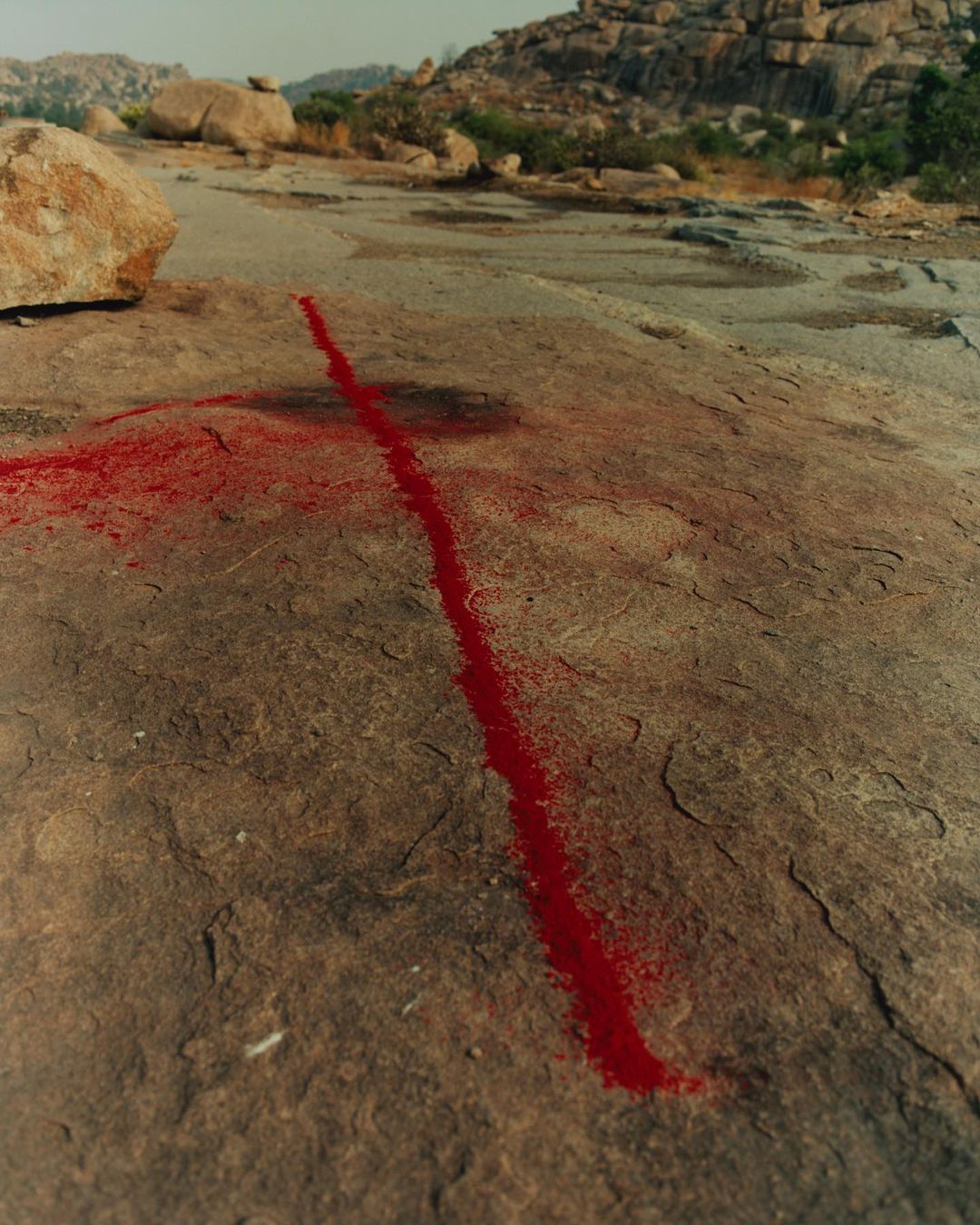 Landscape featuring a red line drawn on a rock formation, with boulders and sparse greenery in the background under a bright sky.