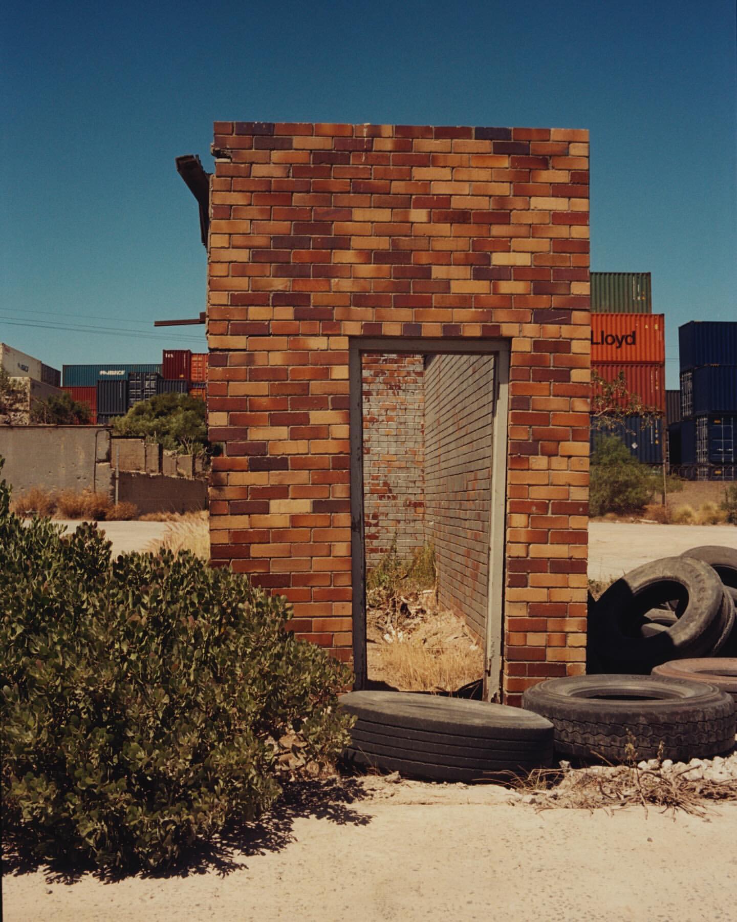Remains of brick wall stand among old tires and vegetation, with shipping containers in the background under a clear blue sky.