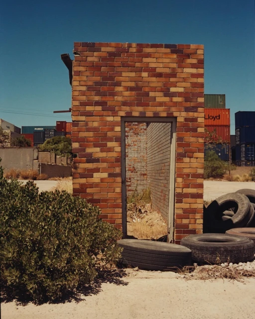 Remains of brick wall stand among old tires and vegetation, with shipping containers in the background under a clear blue sky.