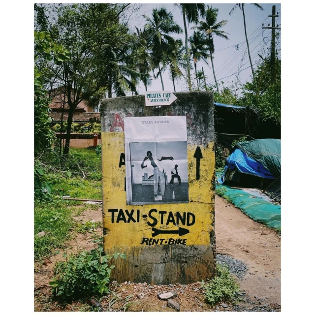 A concrete signpost with hand-painted text and a black and white poster, set in a lush, tropical environment with palm trees and foliage.