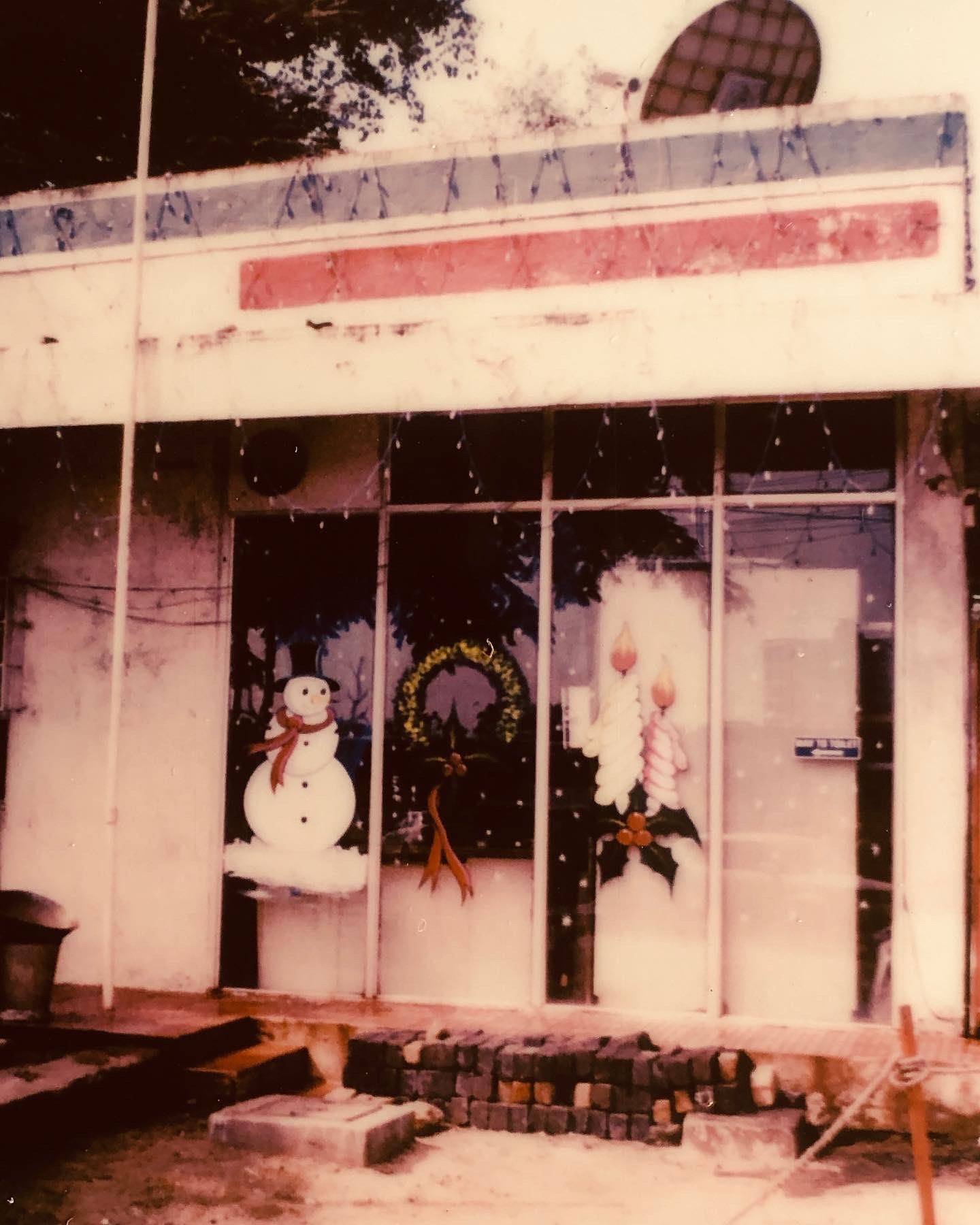Storefront window display featuring painted holiday decorations: a snowman, a Christmas wreath, and two candles with holly.