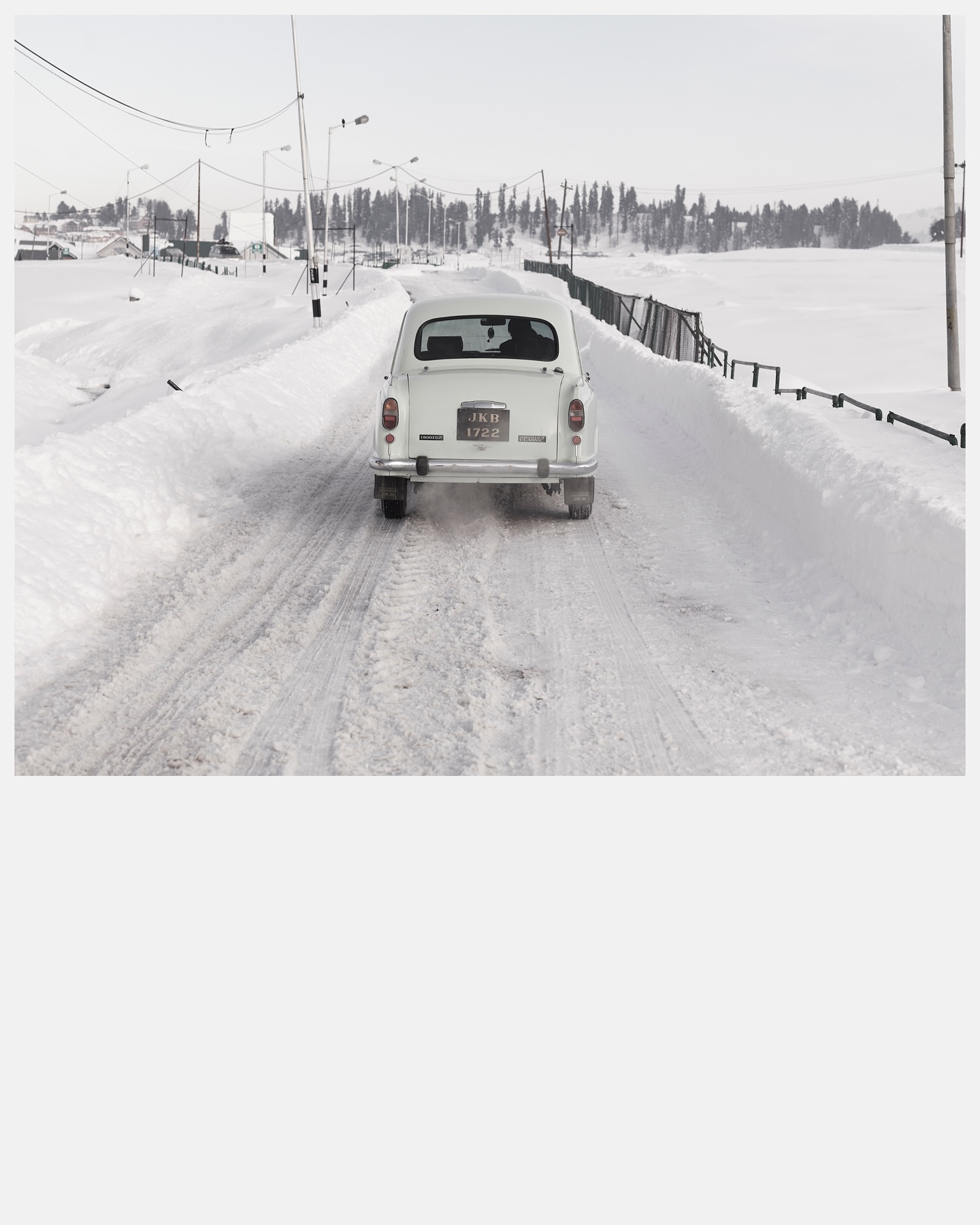 Rear view of a white vintage car driving on a snow-covered road with trees in the background during winter under a clear sky.