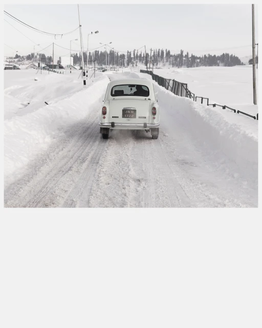 Rear view of a white vintage car driving on a snow-covered road with trees in the background during winter under a clear sky.